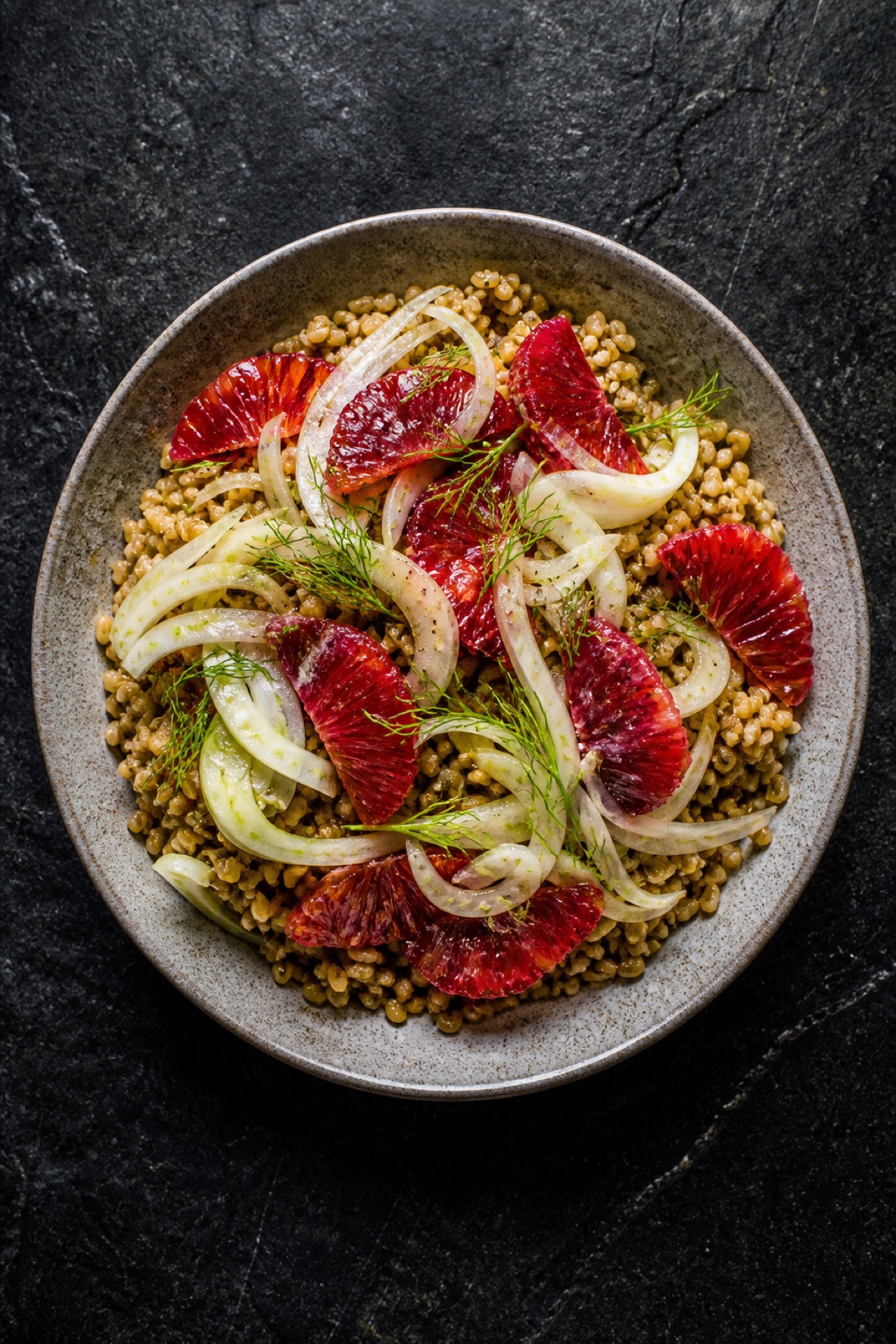 Freekeh grain bowl with shaved fennel and blood orange segments, overhead flat-lay on dark stone, vegan